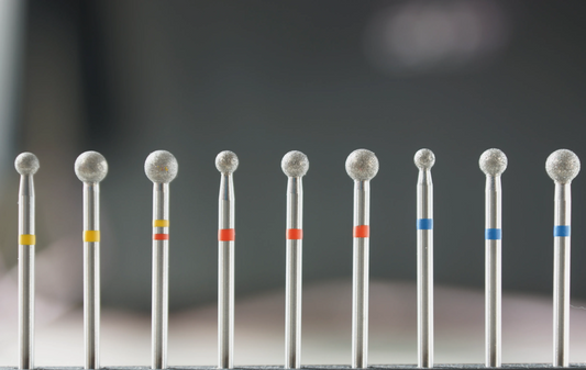 Vue rapprochée d’une série de fraises en diamant de forme boule, aux tailles variées et avec bagues de couleur (jaune, rouge, bleu), utilisées pour le nettoyage des cuticules, la préparation des sillons et le traitement des peaux mortes autour de l’ongle.