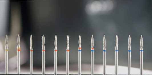 Série de douze fraises en diamant de forme flamme ou effilée, disposées en ligne et différenciées par leurs bagues de couleur (jaune, rouge, bleu), représentant différents niveaux de granulométrie pour les soins des cuticules, callosités et ongles naturels.