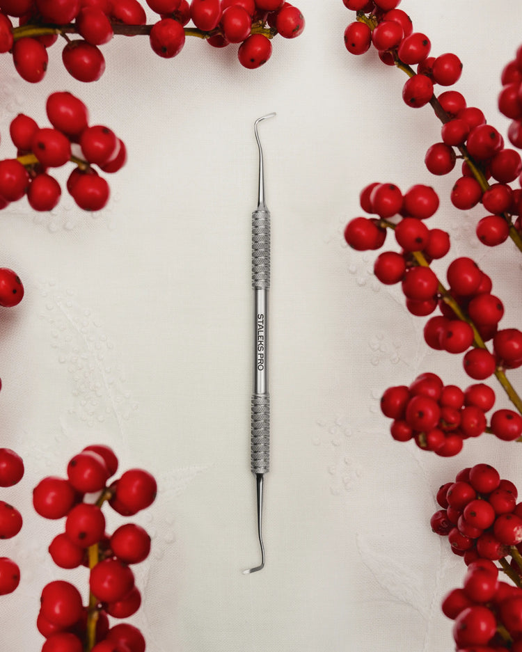 Curette pédicure Staleks Pro en acier inoxydable, posée sur fond blanc décoré de baies rouges, outil professionnel pour le soin des ongles et callosités.