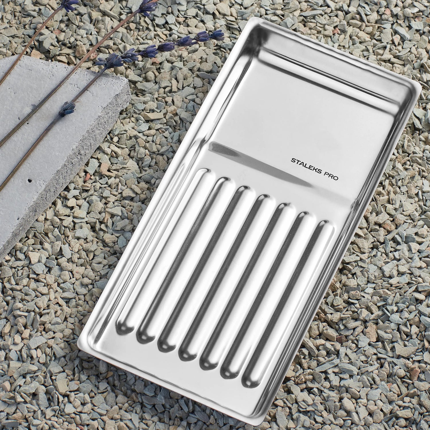Plateau à outils Staleks Pro en acier inoxydable posé sur graviers, accessoire professionnel pour la manucure et la pédicure.