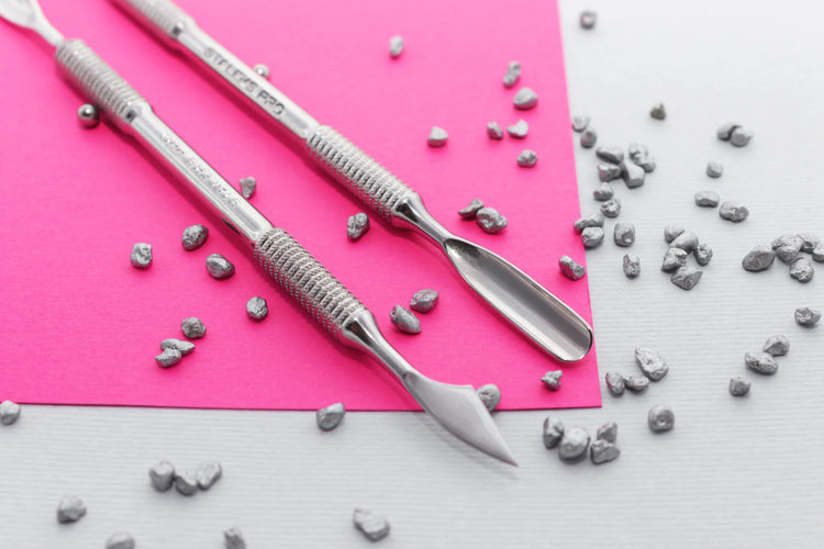 Deux repousse-cuticules Staleks en acier inoxydable posés sur fond rose et blanc, entourés de petits éléments métalliques, outils professionnels de manucure.